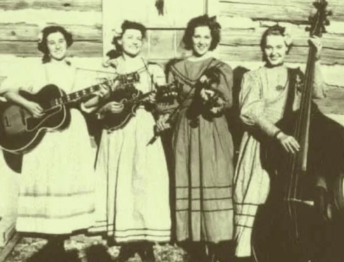 Four women in vintage dresses playing instruments outside a log cabin, smiling and posing for the photo.