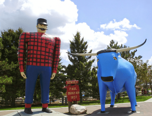 Giant statues of Paul Bunyan and a blue ox in a park, with trees and a sign in the background.