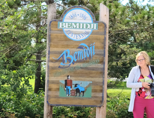 Sign for Bemidji, celebrating its centennial, with a person standing nearby and a dog in the foreground.