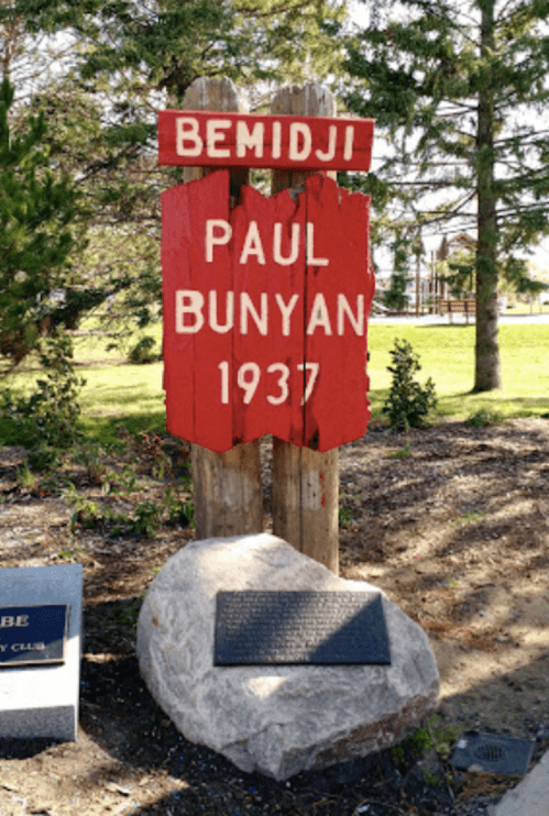 Sign for Paul Bunyan in Bemidji, Minnesota, established in 1937, with a stone base and surrounding greenery.