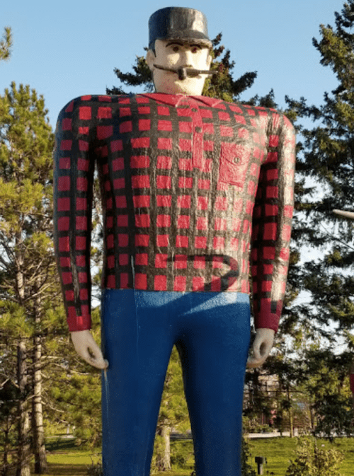 A large wooden statue of a lumberjack in a red and black checkered shirt, blue pants, and a black hat, surrounded by trees.