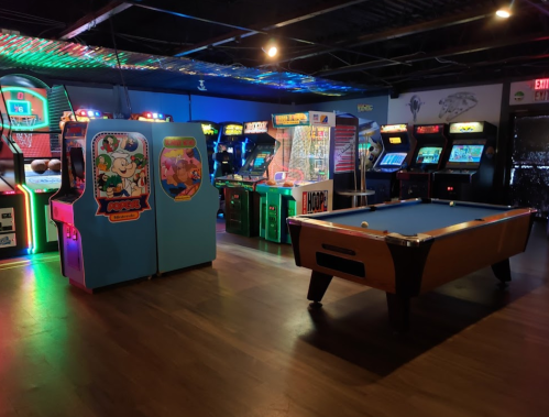 A vibrant arcade with various retro games and a pool table, illuminated by colorful lights.