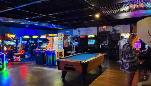 A vibrant arcade with colorful game machines, a pool table, and people enjoying various games in a lively atmosphere.