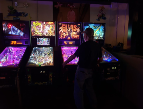 A person playing pinball at an arcade, surrounded by colorful machines featuring various themes.