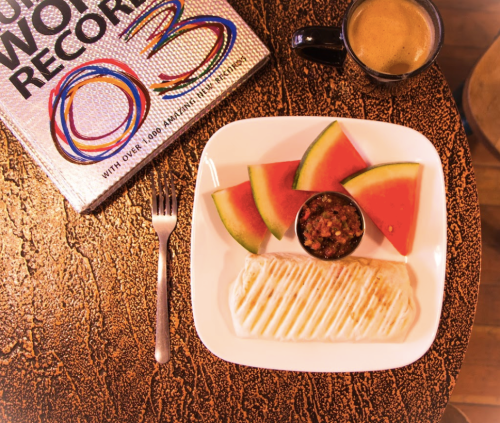 A plate with a burrito, watermelon slices, and salsa, alongside a cup of coffee and a colorful book.