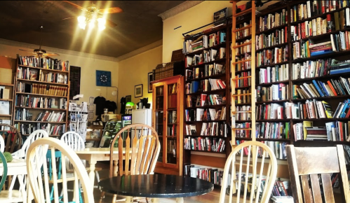 Cozy bookstore interior with wooden tables, chairs, and shelves filled with books. Warm lighting creates a welcoming atmosphere.