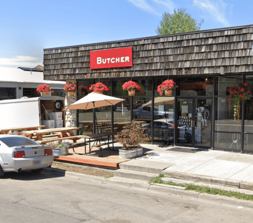 A butcher shop with a wooden exterior, outdoor seating, and flower planters on a sunny street.