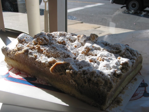A large piece of crumb cake dusted with powdered sugar, sitting on a white paper near a window.