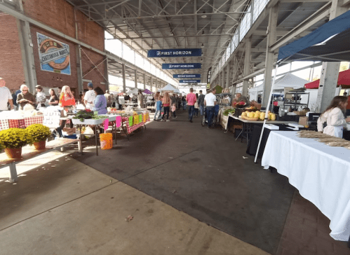 A bustling farmers market with vendors, shoppers, and colorful produce under a covered area.