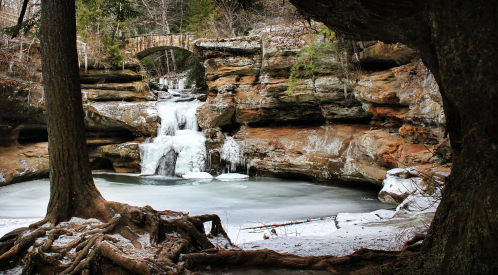 A serene winter scene featuring a frozen waterfall and a stone bridge surrounded by snow-covered rocks and trees.