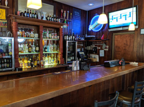 A wooden bar with a variety of liquor bottles, taps, and a TV displaying a crossword puzzle in a cozy setting.