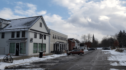 A snowy street scene featuring white buildings, parked cars, and a church steeple in the background under a cloudy sky.