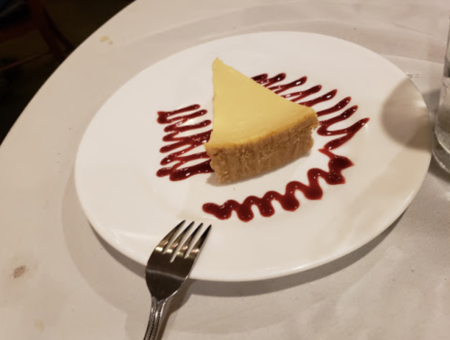 A slice of cheesecake on a white plate, drizzled with raspberry sauce, with a fork beside it.