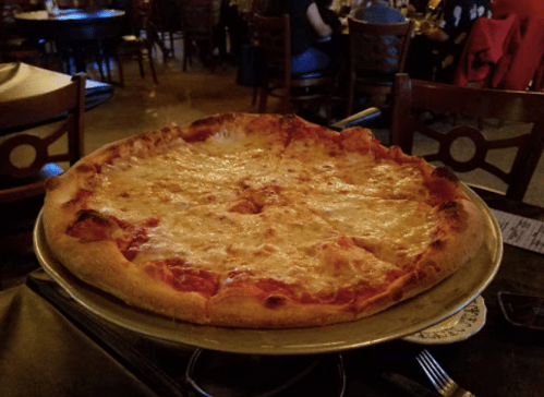 A large cheese pizza on a metal tray, with a blurred restaurant background.