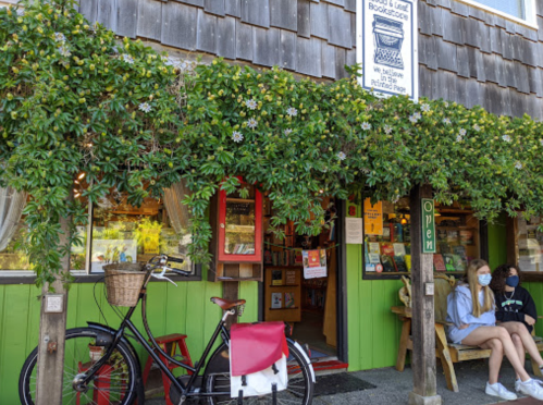A quaint bookstore with a green facade, bike outside, and two people sitting on benches, surrounded by greenery.