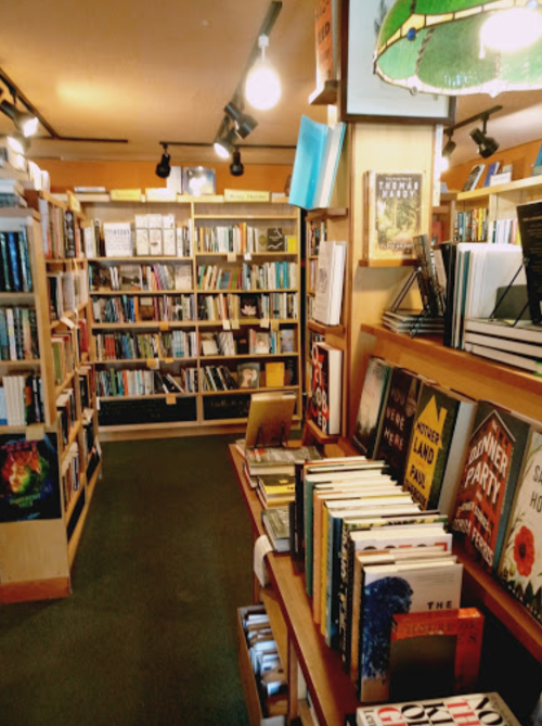 A cozy bookstore aisle lined with shelves of books and a display of titles on wooden tables. Warm lighting enhances the atmosphere.