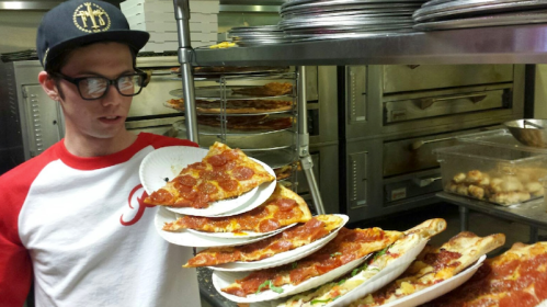 A person in a baseball cap and glasses holds multiple plates of pepperoni pizza in a kitchen setting.