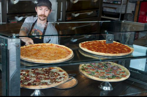 A man with a mustache serves pizza from a glass display case featuring various pizza types.