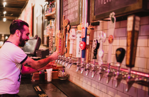 A bartender pours a drink from a row of beer taps in a lively bar setting.