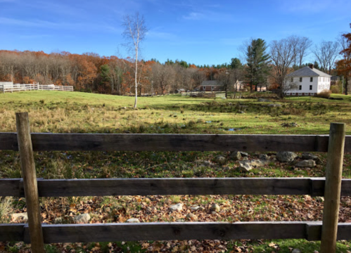 A wooden fence in the foreground with a scenic view of a field, trees, and a house in the background on a clear day.