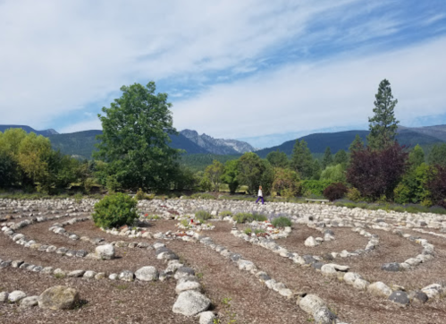 A stone labyrinth surrounded by trees and mountains under a blue sky with scattered clouds.