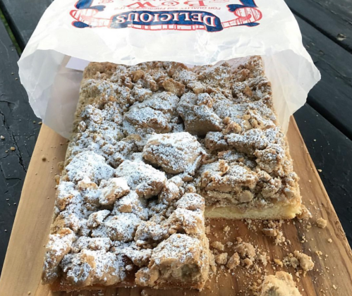 A rectangular piece of crumb cake topped with powdered sugar, partially sliced, on a wooden board.