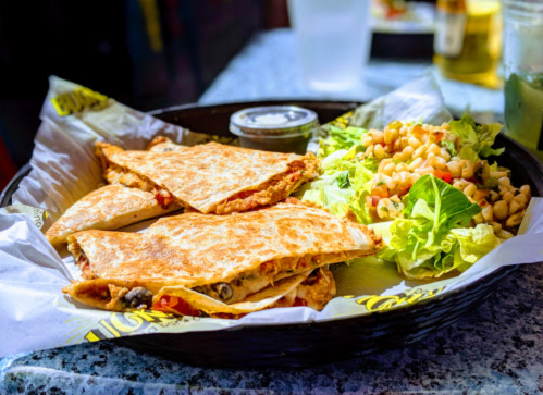 A plate of quesadillas with a side of salad and dipping sauce, served in a black basket on a table.