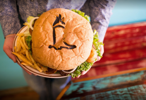 A person holds a large burger with a unique design on the bun, accompanied by a side of fries.
