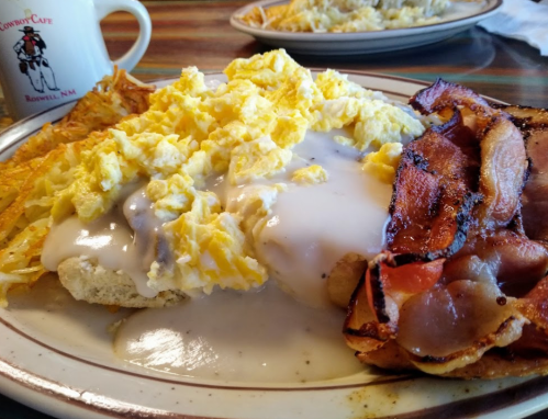 A plate of scrambled eggs with gravy, hash browns, and crispy bacon, served in a diner setting.