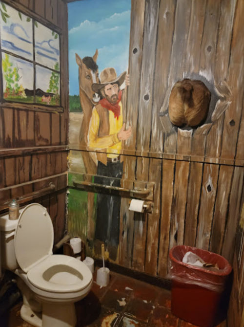 A rustic bathroom with a mural of a cowboy and horse on the wall, featuring a toilet and trash can in the foreground.