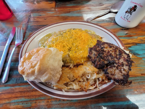 A plate of food featuring green chili cheese, grilled meat, hash browns, and a biscuit on a wooden table.