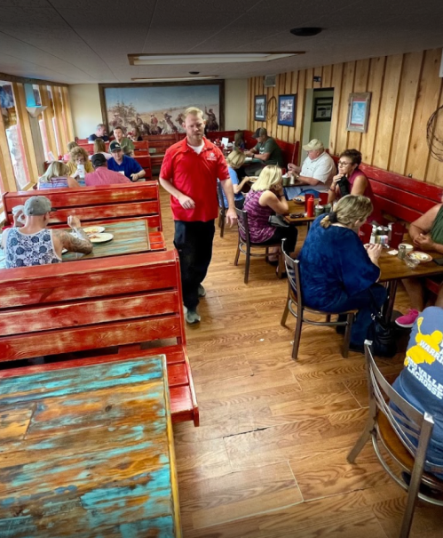 A busy restaurant interior with diners at wooden tables and a server walking through. Warm, rustic decor.