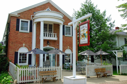 A charming brick building with a white porch, outdoor seating, and a sign for a restaurant. Green trees surround the area.