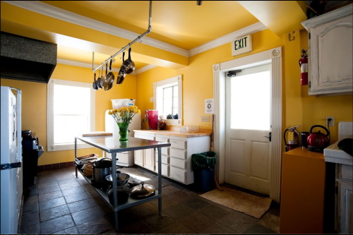 Bright yellow kitchen with a central island, hanging pots, and a door leading outside. Windows provide natural light.