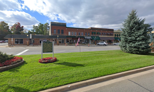 A street view of a small town with shops, green grass, and a sign, under a partly cloudy sky.