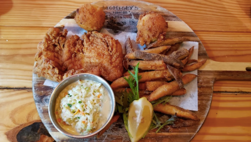 A wooden platter with fried chicken, coleslaw, sweet potato fries, and two hush puppies, garnished with a lemon wedge.