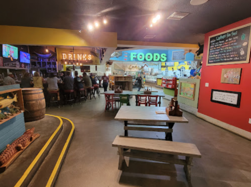 A vibrant restaurant interior with a bar, colorful signs for "Drinks" and "Foods," and wooden tables.