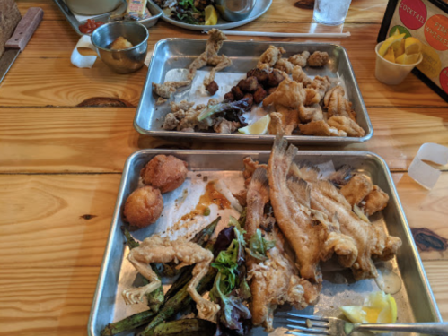 Two metal trays filled with various fried fish, chicken, and sides, garnished with lemon and greens on a wooden table.