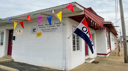 A storefront with colorful flags and an "Open" sign, featuring a sign for a shop selling rocks, minerals, and jewelry.