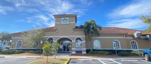 A large, tan building with a tile roof and palm trees, featuring a sign and people entering. Clear blue sky above.