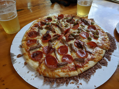 A pizza topped with pepperoni, sausage, and jalapeños, served on a cardboard base with two glasses of beer nearby.