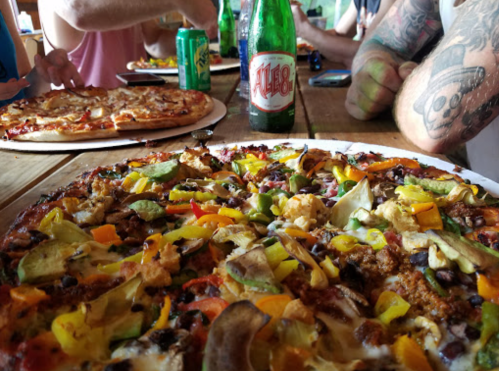 A close-up of a colorful veggie pizza with various toppings, surrounded by drinks and another pizza in the background.