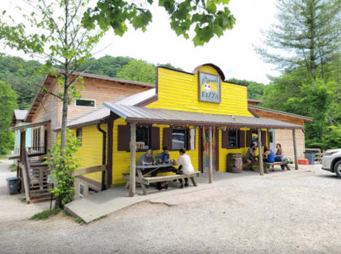 A colorful yellow restaurant with outdoor seating, surrounded by trees and mountains, on a gravel path.