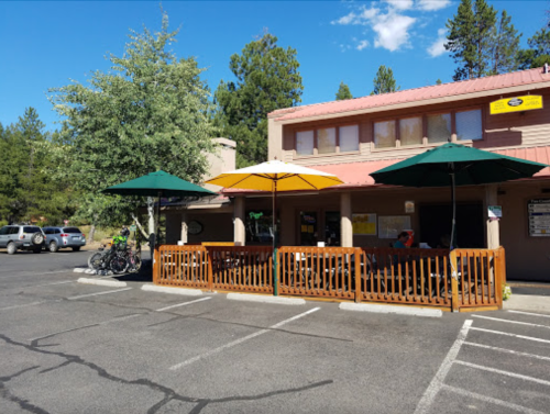 A restaurant with outdoor seating under colorful umbrellas, surrounded by trees and parked cars in a sunny area.