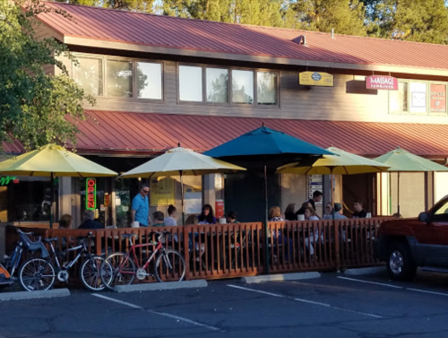 Outdoor dining area with umbrellas, people seated at tables, and bicycles parked nearby in a cozy restaurant setting.