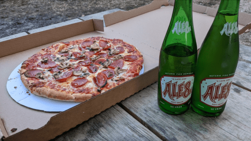 A pizza with pepperoni and mushrooms next to two bottles of Ale-8 soda on a wooden table.
