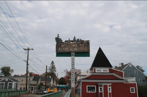 Sign welcoming visitors to Kennebunkport, with a cloudy sky and buildings in the background.