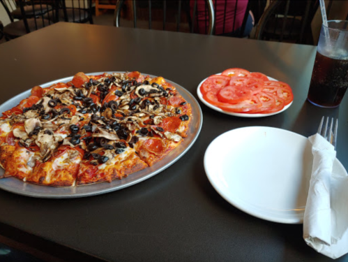 A pizza topped with mushrooms, olives, and pepperoni on a metal tray, alongside a plate of sliced tomatoes and a drink.