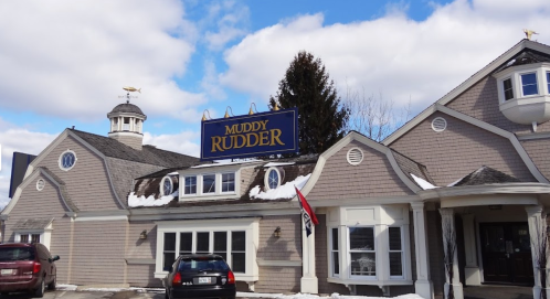 Exterior of the Muddy Rudder restaurant, featuring a large sign and a snow-covered roof under a cloudy sky.