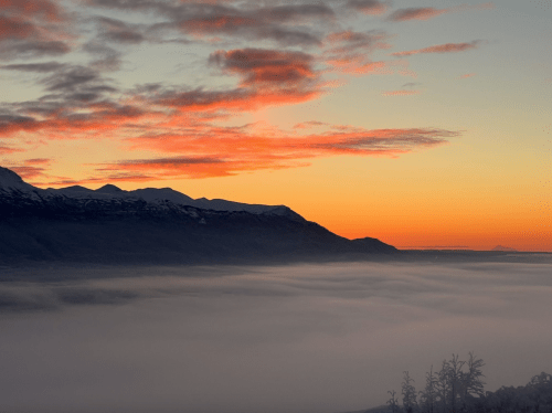 A serene landscape at sunset, with mountains silhouetted against a colorful sky and a blanket of fog below.